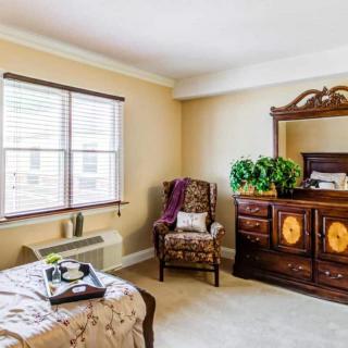 A cozy bedroom with elegant wooden furniture, featuring a bed, armchair, and dresser near a window.