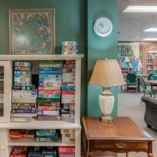 A cozy living room with a puzzle-filled bookshelf, a clock on the wall, and a lamp on a wooden side table.