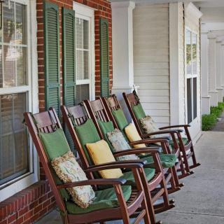 A row of wooden rocking chairs with cushions line a porch outside a brick house.