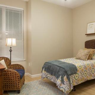A cozy bedroom with a single bed, wicker chair, and a standing lamp beside a window with blinds.