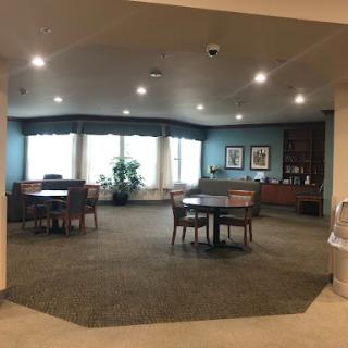 A cozy lounge area featuring tables and chairs, illuminated by soft lighting, with large windows letting in natural light.