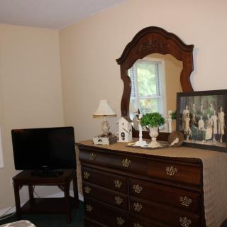 A cozy bedroom corner featuring a traditional wooden dresser with a mirror, decorated with lamps and plants, alongside a TV on a stand.
