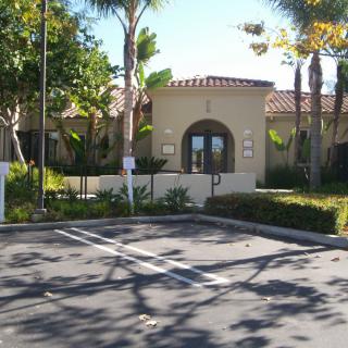 A sunny view of a building entrance with a parking lot and surrounding trees.