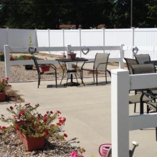 A sunny outdoor patio area with tables and chairs, surrounded by a white fence and decorated with potted flowers.
