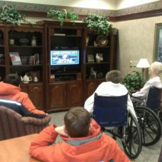 A group of people, including individuals in wheelchairs, gathered around a television in a living room setting.