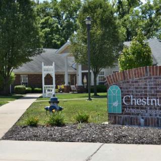 A residential complex with a brick sign in the foreground and a well-maintained landscape featuring trees and pathways.