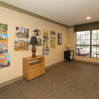 A well-lit room with artwork on the walls, a wooden cabinet with a lamp, and a large window.