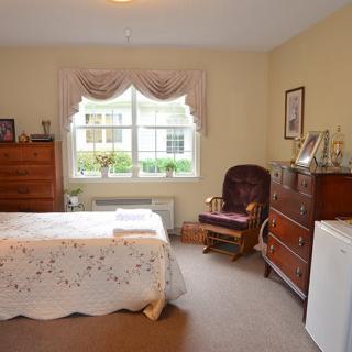 A cozy bedroom featuring a bed with a lace bedspread, a wooden dresser and chest of drawers, a rocking chair, and a window with draped curtains.