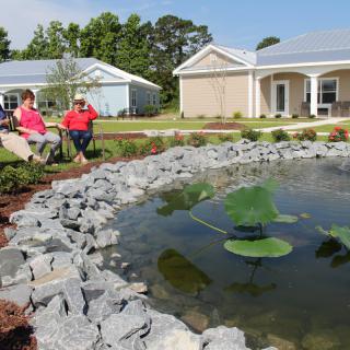 Two people sitting by a pond in a landscaped garden with houses in the background.