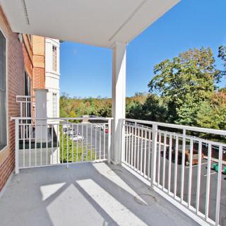 A sunny balcony with white railings overlooking a parking area and trees.
