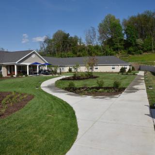 A single-story house with a curved pathway leading through a landscaped garden area with trees in the background under a clear blue sky.