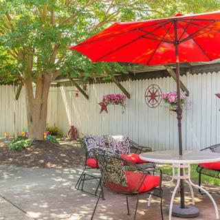 A cozy patio area with red-cushioned chairs under a red umbrella, surrounded by a landscaped garden and white fence.