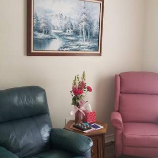 A cozy living room corner with two recliner chairs, a side table adorned with flowers, and a serene landscape painting on the wall.