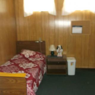 A small bedroom with wood-paneled walls, featuring a single bed with floral bedding, a side table, and a lamp.