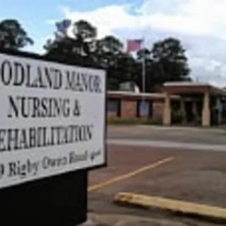 Entrance sign for Woodland Manor Nursing & Rehabilitation with the building and a flag in the background.