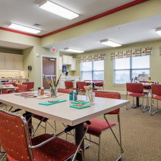 A cozy dining room with tables and chairs, featuring a kitchen area and large windows.