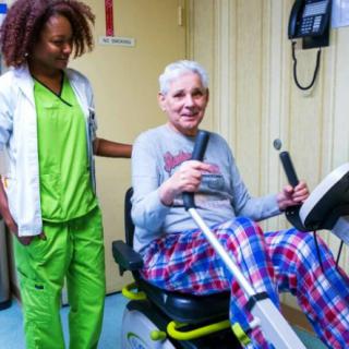 An elderly person using exercise equipment with the assistance of a healthcare worker.
