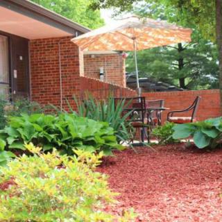 A cozy patio area with a table and chairs under an umbrella, surrounded by lush plants and mulch.