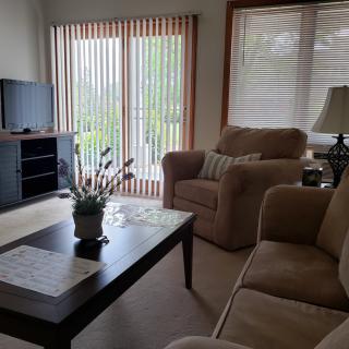 A cozy living room with a sofa set, coffee table, and television, featuring large windows with blinds.