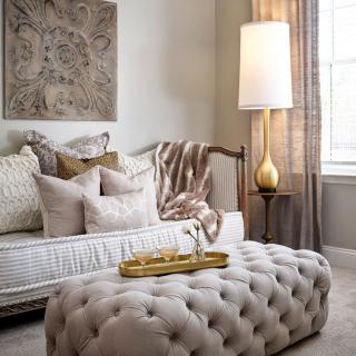 A cozy living room with neutral tones, featuring a stylish lamp, tufted ottoman, and decorative pillows.