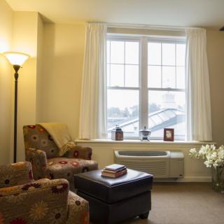 A cozy living room interior with an armchair, ottoman, and a large window letting in natural light.