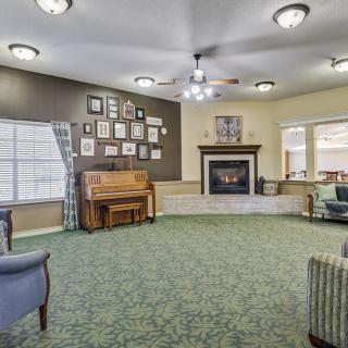 A cozy living room featuring a piano, fireplace, and seating area with green carpet and framed pictures on the wall.