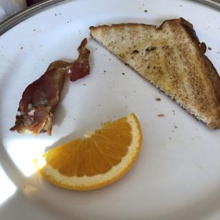 A breakfast plate with a slice of toast, a strip of bacon, and an orange slice.