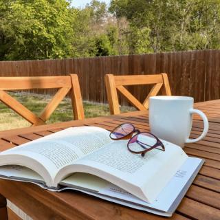 A relaxing outdoor reading setup with an open book, glasses, and a coffee cup on a wooden table.