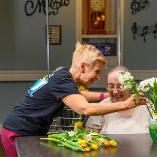 A caregiver joyfully interacts with an elderly person while arranging flowers, showcasing kindness and companionship.