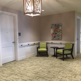 A cozy seating area with two green chairs and a small table set against a beige wall, adorned with artwork and illuminated by an overhead lamp.