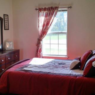 A cozy bedroom with a red bedspread, wooden dresser, and sunlight streaming through the window.