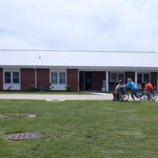 People in wheelchairs are being assisted on a sidewalk outside a brick building.