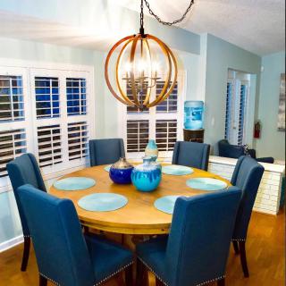 A cozy dining room with a round wooden table, surrounded by blue upholstered chairs and illuminated by a modern chandelier.