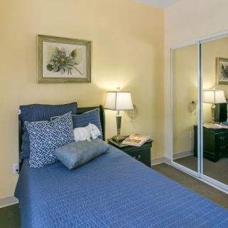 A cozy bedroom with a single bed dressed in blue bedding, accompanied by a nightstand and decorative lamps. A mirrored closet reflects the warm tones of the room.