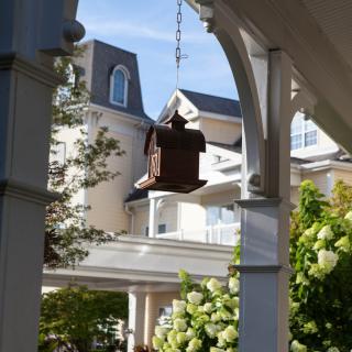 A wooden birdhouse hangs in a garden with a large house in the background, surrounded by greenery and bathed in sunlight.