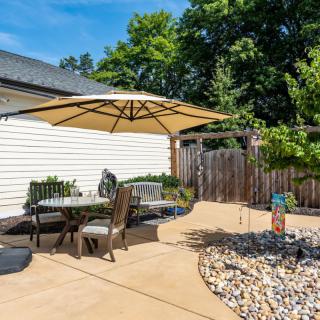 A sunny backyard patio with outdoor furniture and an umbrella, surrounded by a garden and fence.