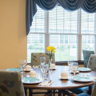 A neatly set dining table with flowers by a large window.