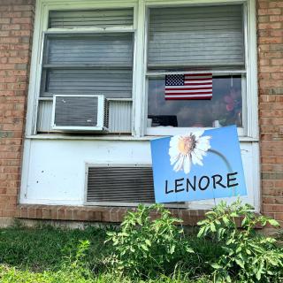 A window with an air conditioner and an American flag, below which is a sign featuring a flower and the name 'Lenore', set against a brick wall and surrounded by grass.