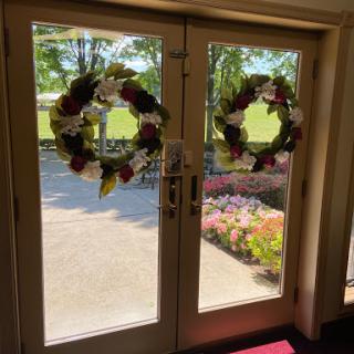 A sunny entrance featuring elegant floral wreaths on double doors, with a glimpse of the beautiful outdoor scenery.