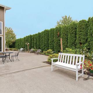 A neat patio area with a white bench, table, and chairs, bordered by a tall green hedge.