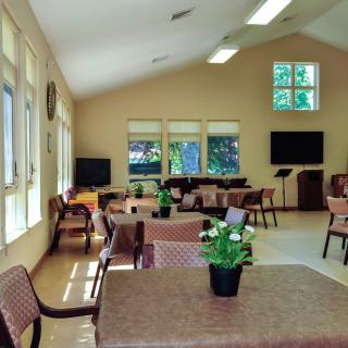 A spacious community room with tables and chairs, decorated with potted plants and natural light.