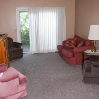A cozy living room with sofas, armchairs, and a lamp, featuring carpeted flooring and large windows with curtains.