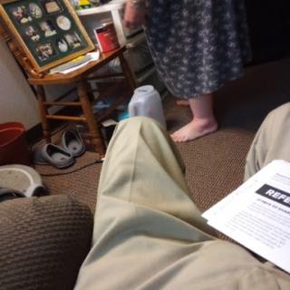 A casual moment in a living room, with a person standing and interacting with their surroundings while another individual relaxes on the couch.