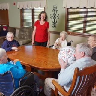 A group of elderly people are sitting around a table in a nursing home, participating in an activity with assistance from a caregiver.