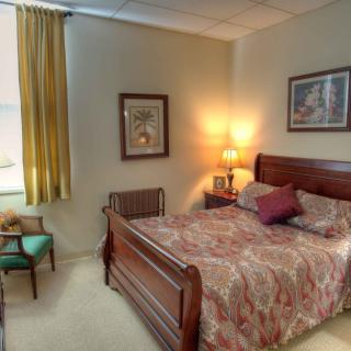 A cozy bedroom featuring a wooden bed, bedside lamp, artwork on the walls, and natural light filtering through curtains.