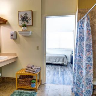 A view of a bathroom leading into a bedroom, featuring a shower curtain, sink, and neatly arranged towels.