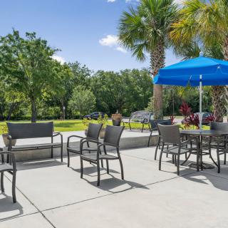 A sunny outdoor patio area with tables, chairs, and a blue umbrella surrounded by lush greenery.
