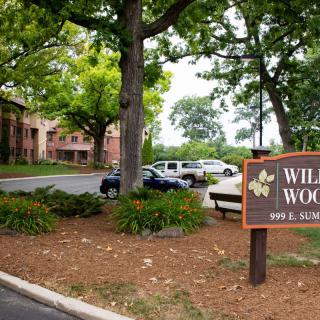 An apartment complex surrounded by trees with a visible parking area and a sign reading 'Willow Woods'.