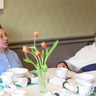 Two elderly individuals are seated at a dining table having a conversation, with a vase of flowers in the center.