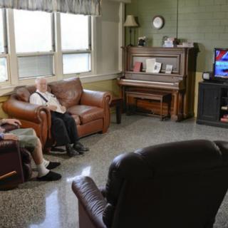 Two seniors sitting in a living room with a piano and watching television.